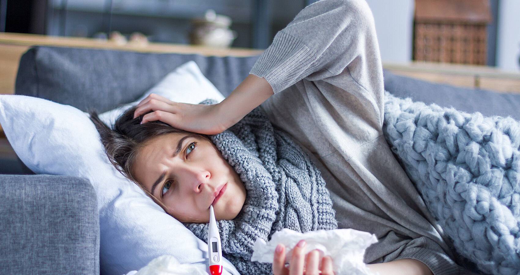 Frau liegt mit einem Fieberthermometer im Mund auf einer Couch